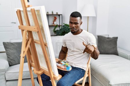 Téléchargez les photos : Jeune homme africain peinture sur toile à la maison pointant vers le bas regardant triste et bouleversé, indiquant la direction avec les doigts, malheureux et déprimé. - en image libre de droit