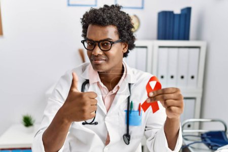 Photo for African doctor man holding support red ribbon smiling happy and positive, thumb up doing excellent and approval sign - Royalty Free Image