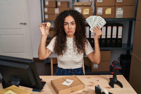 Téléchargez les photos : Young hispanic woman working at small business ecommerce holding money and bitcoin depressed and worry for distress, crying angry and afraid. sad expression. - en image libre de droit