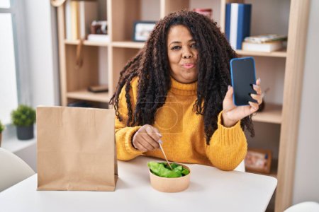 Téléchargez les photos : Plus taille hispanique femme manger emporter salade tenant smartphone souriant regardant vers le côté et regardant loin en pensant. - en image libre de droit