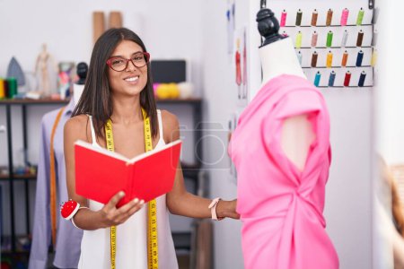 Téléchargez les photos : Young beautiful hispanic woman tailor reading book looking dress at tailor shop - en image libre de droit