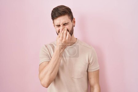 Téléchargez les photos : Hispanic man with beard standing over pink background smelling something stinky and disgusting, intolerable smell, holding breath with fingers on nose. bad smell - en image libre de droit