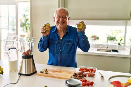Foto de Hombre mayor sonriendo confiado sosteniendo dos aguacate medianos en la cocina - Imagen libre de derechos