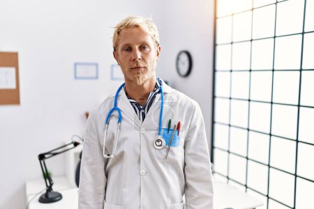 Photo for Young blond man wearing doctor uniform and stethoscope at clinic looking sleepy and tired, exhausted for fatigue and hangover, lazy eyes in the morning. - Royalty Free Image