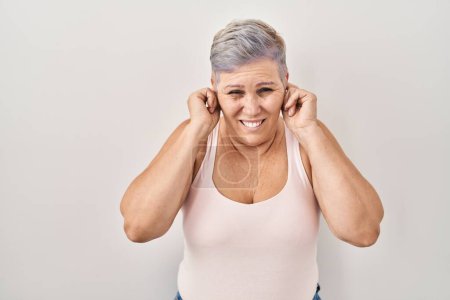 Foto de Middle age caucasian woman standing over white background covering ears with fingers with annoyed expression for the noise of loud music. deaf concept. - Imagen libre de derechos