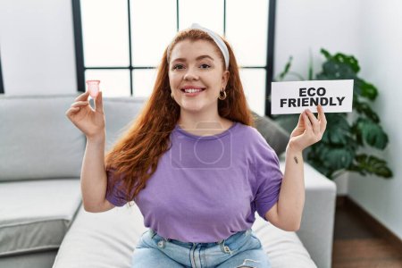 Photo for Young redhead woman smiling confident holding menstrual cup and eco friendly banner at home - Royalty Free Image