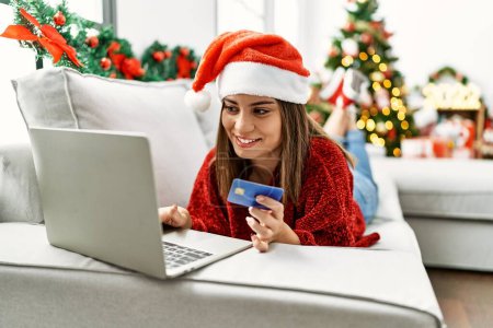 Photo for Young hispanic girl buying using credit card and laptop lying on the sofa by christmas tree at home. - Royalty Free Image