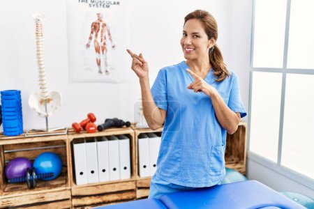 Photo for Middle age hispanic physiotherapist woman working at pain recovery clinic smiling and looking at the camera pointing with two hands and fingers to the side. - Royalty Free Image