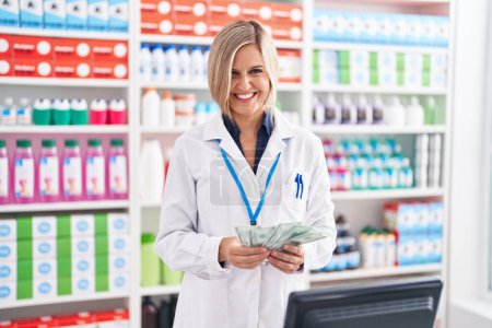 Photo for Young blonde woman pharmacist smiling confident counting dollars at pharmacy - Royalty Free Image