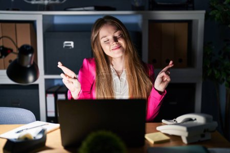 Téléchargez les photos : Young caucasian woman working at the office at night gesturing finger crossed smiling with hope and eyes closed. luck and superstitious concept. - en image libre de droit