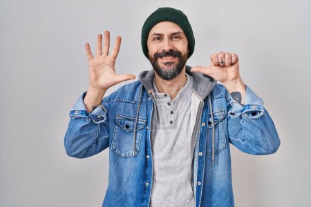 Téléchargez les photos : Jeune homme hispanique avec des tatouages portant une casquette en laine montrant et pointant vers le haut avec les doigts numéro six tout en souriant confiant et heureux. - en image libre de droit
