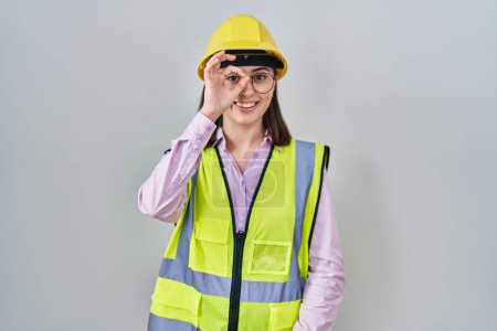 Foto de Chica hispana vistiendo uniforme constructor y hardhat haciendo buen gesto con la mano sonriente, los ojos mirando a través de los dedos con cara feliz. - Imagen libre de derechos