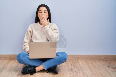 Foto de Mujer joven usando el ordenador portátil sentado en el suelo en casa aburrido bostezo cansado cubriendo la boca con la mano. inquietud y somnolencia. - Imagen libre de derechos