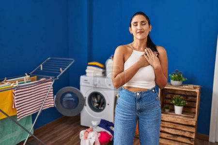 Photo for Young hispanic woman at laundry room smiling with hands on chest with closed eyes and grateful gesture on face. health concept. - Royalty Free Image