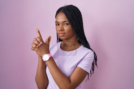 Foto de Mujer afroamericana con trenzas de pie sobre fondo rosa sosteniendo arma simbólica con gesto de mano, jugando a matar armas de fuego, cara enojada - Imagen libre de derechos
