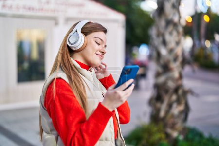 Téléchargez les photos : Jeune femme blonde souriante à l'écoute de musique dans la rue - en image libre de droit