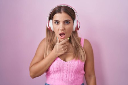 Foto de Young blonde woman listening to music using headphones looking fascinated with disbelief, surprise and amazed expression with hands on chin - Imagen libre de derechos