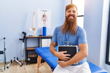 Téléchargez les photos : Jeune homme rousse portant un uniforme de physiothérapeute tenant une mallette à la clinique de physiothérapie - en image libre de droit