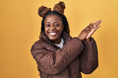 Foto de African woman with braided hair standing over yellow background clapping and applauding happy and joyful, smiling proud hands together - Imagen libre de derechos
