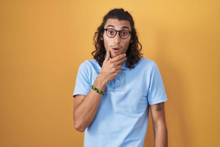 Photo for Young hispanic man standing over yellow background looking fascinated with disbelief, surprise and amazed expression with hands on chin - Royalty Free Image