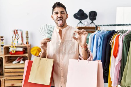Photo for Young hispanic man holding shopping bags, dollars and bitcoin sticking tongue out happy with funny expression. - Royalty Free Image