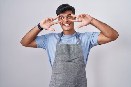 Photo for Hispanic young man wearing apron over white background doing peace symbol with fingers over face, smiling cheerful showing victory - Royalty Free Image