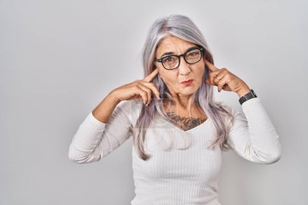 Middle age woman with grey hair standing over white background covering ears with fingers with annoyed expression for the noise of loud music. deaf concept. 