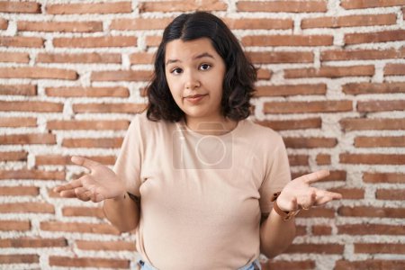 Photo for Young hispanic woman standing over bricks wall clueless and confused with open arms, no idea concept. - Royalty Free Image