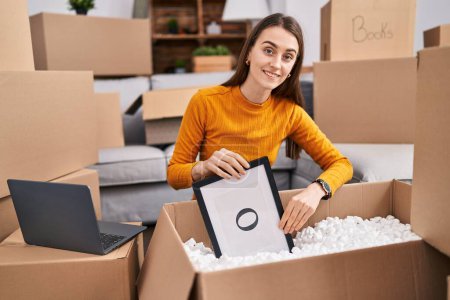 Photo for Young caucasian woman smiling confident unpacking cardboard box at new home - Royalty Free Image