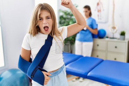 Foto de Blonde little girl wearing arm on sling at rehabilitation clinic annoyed and frustrated shouting with anger, yelling crazy with anger and hand raised - Imagen libre de derechos