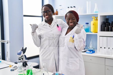 Téléchargez les photos : Deux femmes africaines travaillant au laboratoire scientifique pointant le pouce vers le côté souriant heureux avec la bouche ouverte - en image libre de droit