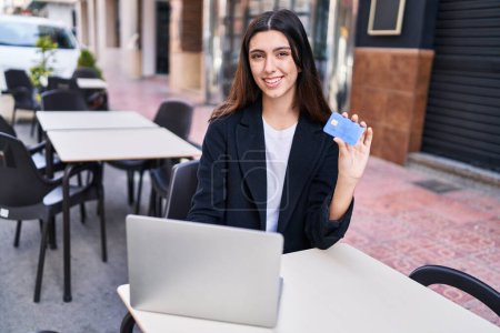 Photo for Young beautiful hispanic woman using laptop and credit card sitting on table at coffee shop terrace - Royalty Free Image