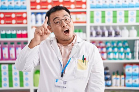 Photo for Chinese young man working at pharmacy drugstore pointing finger up with successful idea. exited and happy. number one. - Royalty Free Image