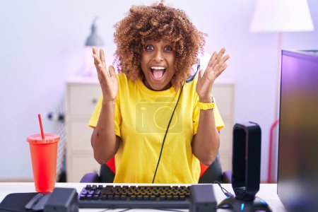 Téléchargez les photos : Jeune femme hispanique aux cheveux bouclés jouant à des jeux vidéo portant des écouteurs célébrant fou et étonné pour le succès avec les bras levés et les yeux ouverts hurlant excités. concept gagnant - en image libre de droit
