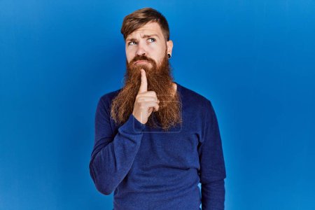 Téléchargez les photos : Homme rousse à longue barbe portant un pull bleu décontracté sur fond bleu pensant concentré sur le doute avec le doigt sur le menton et regardant vers le haut se demandant - en image libre de droit