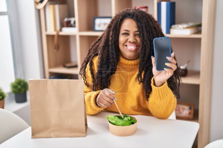 Téléchargez les photos : Plus taille femme hispanique manger emporter salade tenant smartphone souriant avec un sourire heureux et cool sur le visage. montrant les dents. - en image libre de droit