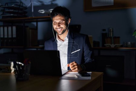 Foto de Hombre latino guapo trabajando en la oficina por la noche luciendo positivo y feliz de pie y sonriendo con una sonrisa confiada mostrando los dientes - Imagen libre de derechos