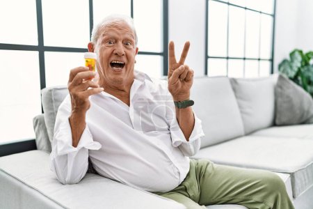 Foto de Hombre mayor sosteniendo pastillas sonriendo con la cara feliz guiñando el ojo a la cámara haciendo señal de victoria con los dedos. número dos. - Imagen libre de derechos