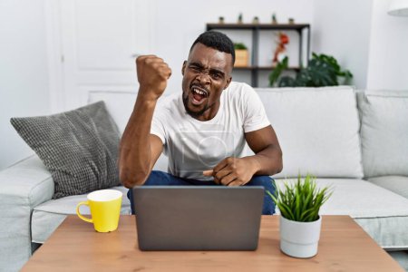 Foto de Hombre africano joven usando el ordenador portátil en casa enojado y loco levantando el puño frustrado y furioso mientras gritaba con ira. rabia y concepto agresivo. - Imagen libre de derechos