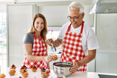 Téléchargez les photos : Couple hispanique moyen âge souriant muffins cuisine heureuse à la cuisine. - en image libre de droit