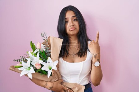 Foto de Mujer morena sosteniendo ramo de flores blancas apuntando hacia arriba mirando triste y molesto, indicando la dirección con los dedos, infeliz y deprimido. - Imagen libre de derechos