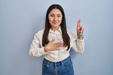 Photo for Young latin woman standing over blue background smiling swearing with hand on chest and fingers up, making a loyalty promise oath - Royalty Free Image