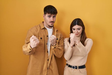Foto de Young hispanic couple standing over yellow background ready to fight with fist defense gesture, angry and upset face, afraid of problem - Imagen libre de derechos