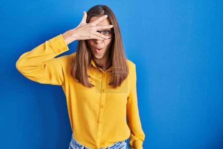 Photo for Young woman wearing glasses standing over blue background peeking in shock covering face and eyes with hand, looking through fingers with embarrassed expression. - Royalty Free Image