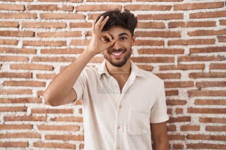 Foto de Hombre árabe con barba de pie sobre ladrillos fondo de la pared haciendo buen gesto con la mano sonriente, los ojos mirando a través de los dedos con cara feliz. - Imagen libre de derechos