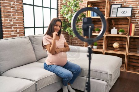 Photo for Young pregnant woman recording vlog tutorial with smartphone at home serious face thinking about question with hand on chin, thoughtful about confusing idea - Royalty Free Image