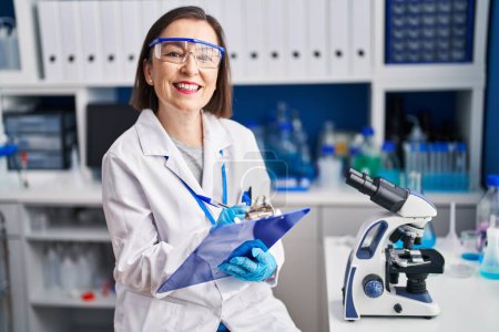 Foto de Middle age woman scientist smiling confident writing on clipboard at laboratory - Imagen libre de derechos