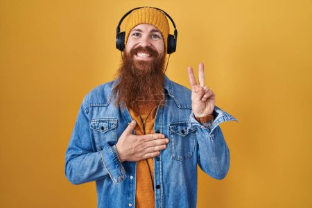 Foto de Hombre caucásico con barba larga escuchando música usando auriculares sonriendo jurando con la mano en el pecho y los dedos hacia arriba, haciendo un juramento de lealtad promesa - Imagen libre de derechos