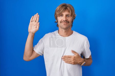 Foto de Hombre de mediana edad de pie sobre fondo azul jurando con la mano en el pecho y la palma abierta, haciendo una promesa de lealtad juramento - Imagen libre de derechos