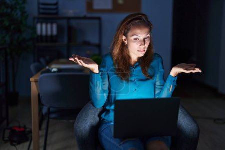 Photo for Brunette woman working at the office at night clueless and confused expression with arms and hands raised. doubt concept. - Royalty Free Image
